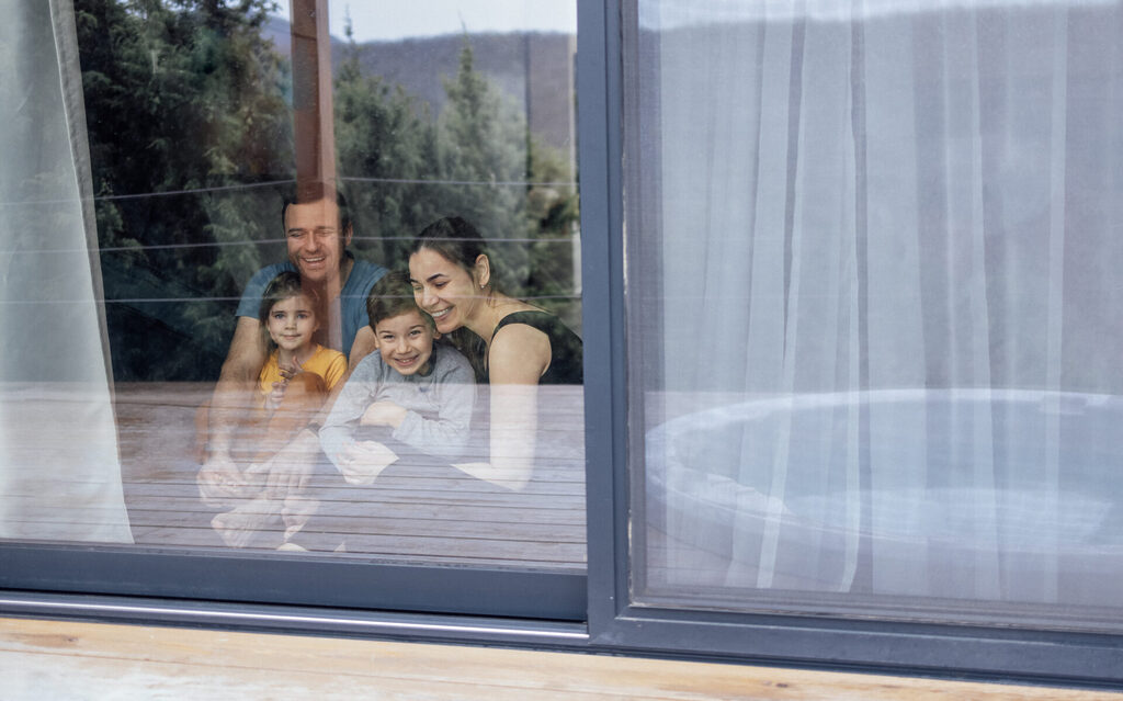 Happy young family is sitting on living room floor Parents and children hug and have fun at home by glass door | GFD Homes benefits of triple glazing: sliding door with family sat down smiling in view.