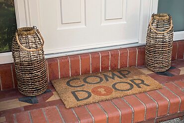 Two tall woven wicker lanterns flank a white front door on a brick stoop, with a coir doormat reading 'WELCOME'.