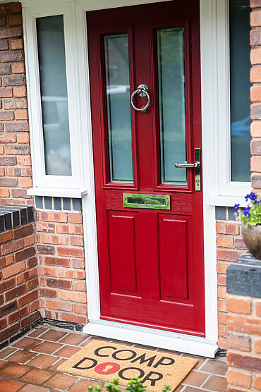 Red front door with glass side panels, silver ring door knocker, and a brick porch.