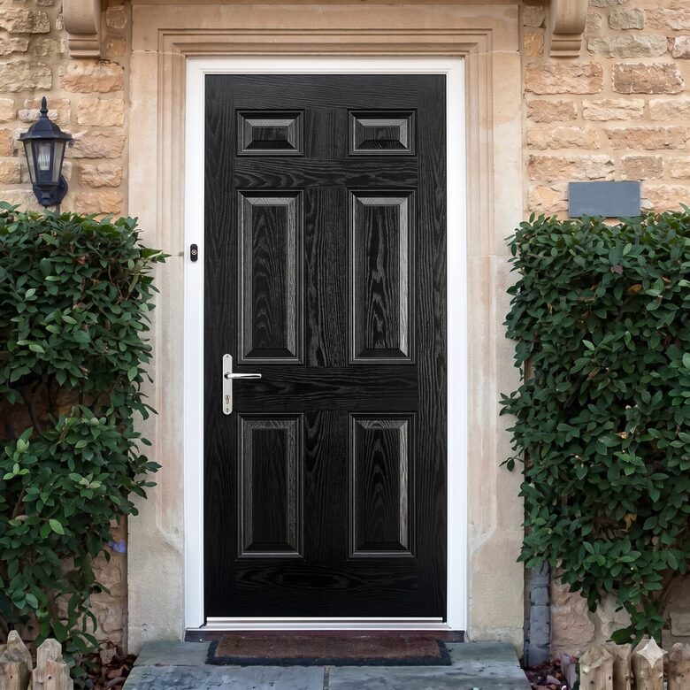 Black front door with decorative panels set in a light stone surround, framed by green shrubs and a lantern to the left.