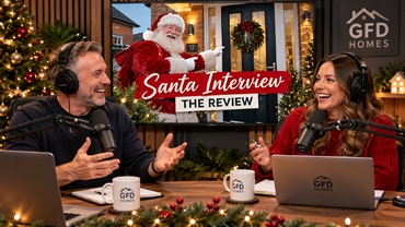 Two hosts in a podcast studio laugh and talk, wearing headphones with microphones, Christmas decor and a TV screen showing 'Santa Interview - The Review' behind them.